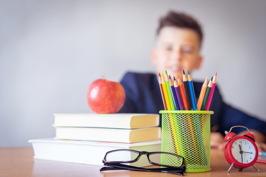 pexels-photo-2781814-2781814 Schoolboy smiling behind a desk with books, pencils, and an alarm clock symbolizing study and creativity.